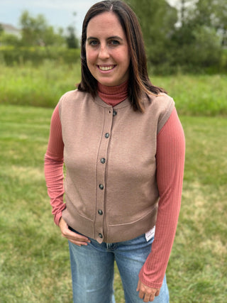 A woman standing outdoors in a sleeveless mocha knit button-front vest over a pink ribbed turtleneck, paired with blue jeans.