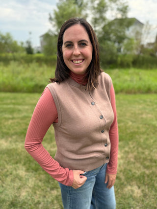 A woman smiling outdoors in a taupe knit button-front vest over a pink ribbed long-sleeve top and blue jeans, standing on a grassy lawn with trees in the background.