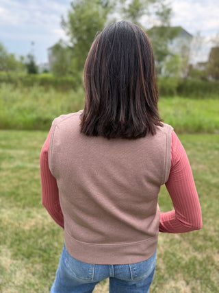 A woman viewed from behind wearing a sleeveless taupe vest over a pink ribbed long-sleeve top and blue jeans.