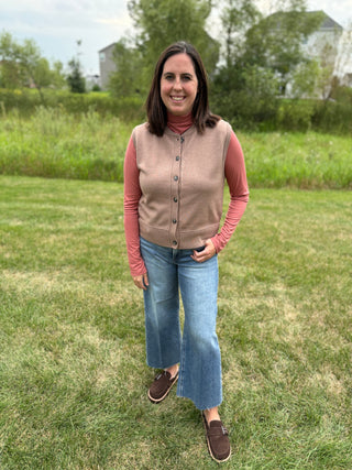 A woman wearing a taupe knit button-up vest over a dusty rose long-sleeve turtleneck, blue wide-leg jeans, and brown slip-on shoes.