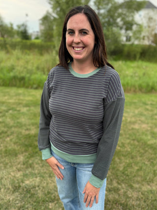 A woman standing outdoors on a grassy field wearing a dark gray and white striped long-sleeve shirt with sage green ribbed trim, paired with light blue jeans.