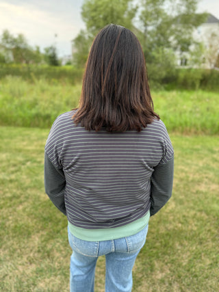 A person viewed from behind wearing a dark gray and white striped long-sleeve top with sage green band on wrist and waist, and light blue jeans.