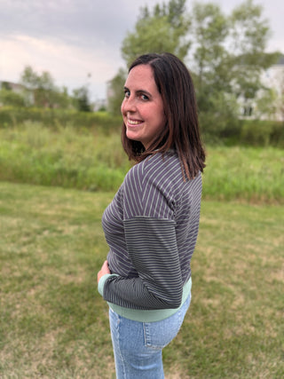 A woman wearing a gray and white vertically striped long-sleeve top with a sage green band on wrist and waist and light blue jeans.