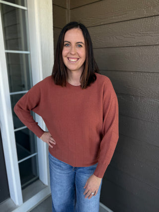 A woman wearing a rust-colored long-sleeve knit sweater and blue jeans.