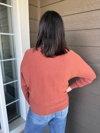 A woman viewed from behind wearing a textured, long-sleeve knit sweater in a rust color, paired with blue jeans.
