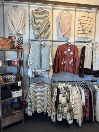 A retail clothing display featuring multiple sweaters and outerwear garments hanging on racks and hooks against a white wall. Visible items include striped knit sweaters, quilted vests, a rust-colored floral cardigan, plaid shirts, and several handbags on a shelving unit to the left.