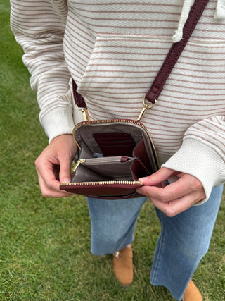 A person outdoors on grass holding open the Payton crossbody bag in the color plum with gold-tone hardware.