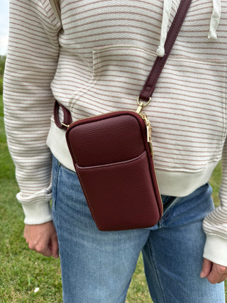 A person wearing a cream and beige striped hoodie and blue jeans carries a plum crossbody phone pouch with gold-tone hardware.