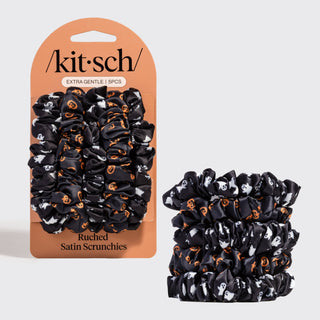 A 5-pack of Kit Sch satin scrunchies in a black, white, and orange Halloween pattern, shown both in their retail packaging and as a loose stack on a light gray background.