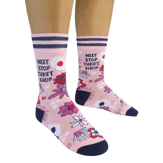 Pink crew socks with a floral pattern and the text 'NEXT STOP THRIFT SHOP' printed on them, shown on a person's feet against a white background.