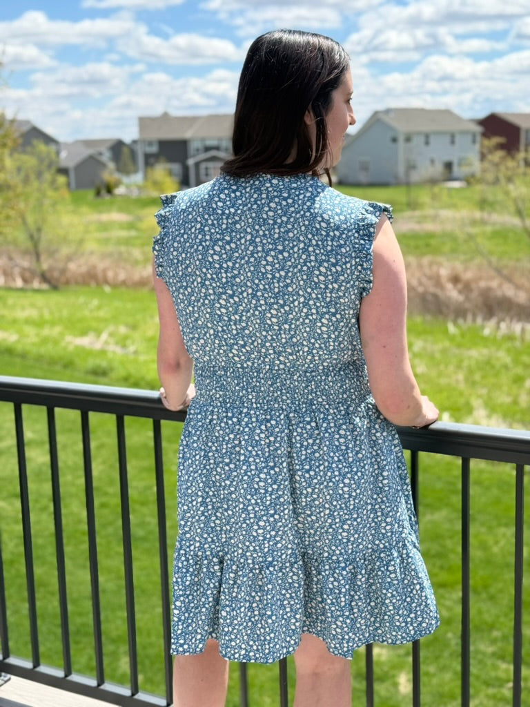 Back view of woman wearing dusty teal ditsy floral mini dress with ruffle cap sleeves and smocked waist
