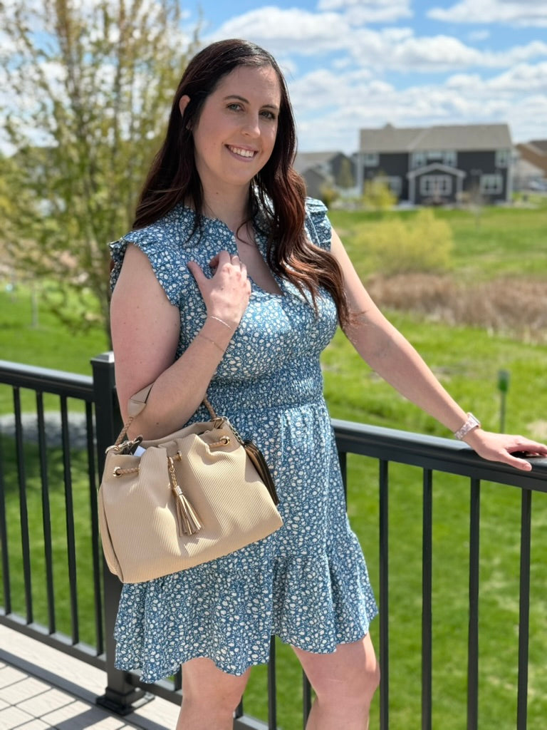 Woman in a blue floral dress holding a beige handbag, standing outdoors with greenery and buildings in the background.