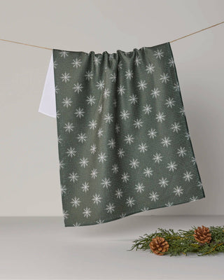 Geometry Snowy tea towel in Juniper with white snowflakes on deep green fabric, hanging above pinecones and greenery