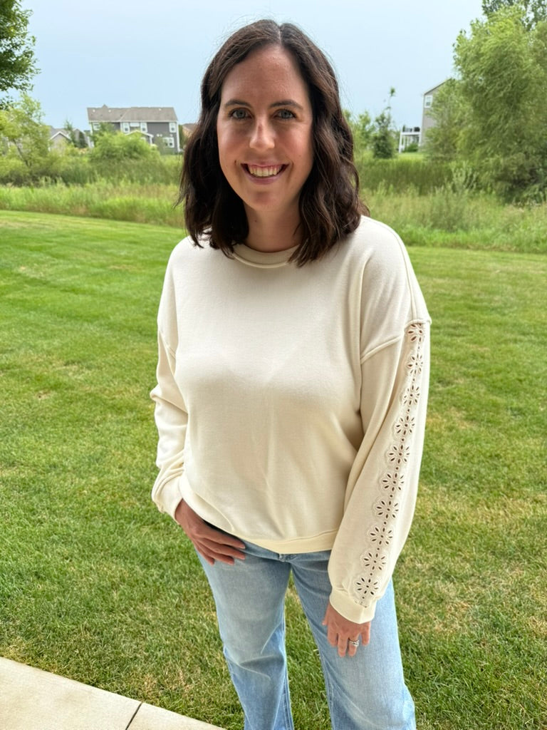 A woman wearing a cream-colored crewneck sweatshirt with decorative cutout detailing on the sleeves, paired with light blue jeans, standing outdoors on a green lawn.