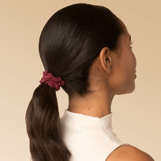 A woman shown from the back and side profile wearing a dark brown hair extension or wig tied back in a ponytail with a textured burgundy scrunchie, against a solid beige background.
