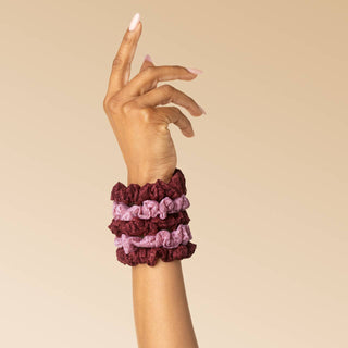 Three fabric scrunchies in burgundy, dusty pink, and mauve are stacked on a person's wrist against a solid beige background.