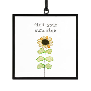 Find Your Sunshine Suncatcher by DEMDACO — a hand-crafted sea glass sunflower with golden yellow and cream petals, dark pebble center, and three pairs of bright green sea glass leaves on a wire stem, inside a square black metal floating frame with "find your sunshine" in typewriter font, hung by a black ribbon

