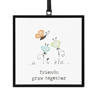 Friends Grow Together Suncatcher by DEMDACO — a whimsical peach sea glass butterfly hovering above two sea glass flowers in soft aqua and pale green on curly wire stems in a hand-drawn garden, inside a square black metal floating frame with "friends grow together" in typewriter font, hung by a black ribbon