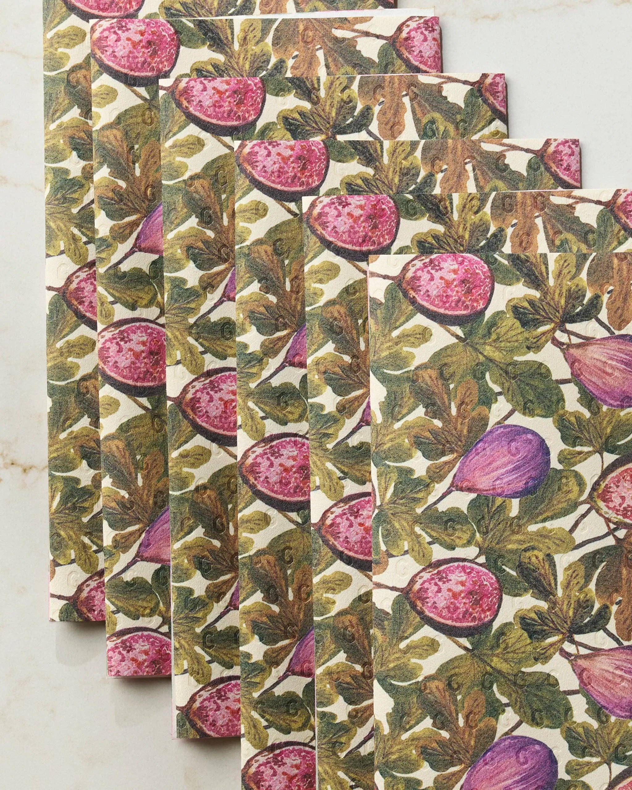 Close-up of Geometry Wild Fig Not Paper Towel set of 6 on white marble showing detailed botanical illustration of sliced pink figs and whole purple figs among earthy green leaves on Geoweave™ fabric