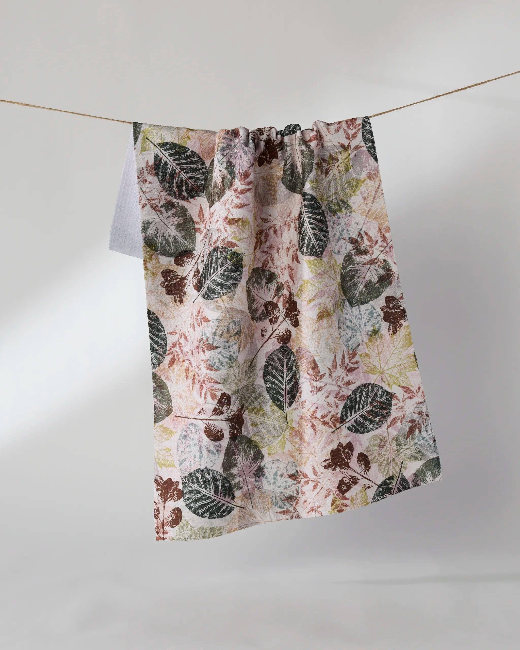 Geometry Woodland Press Tea Towel hanging on a clothesline showing full 18x30 eco-friendly kitchen towel with layered botanical leaf press print in forest green, earthy brown, and dusty rose on blush background
