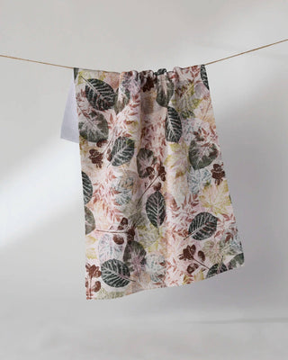 Geometry Woodland Press Tea Towel hanging on a clothesline showing full 18x30 eco-friendly kitchen towel with layered botanical leaf press print in forest green, earthy brown, and dusty rose on blush background
