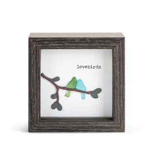 A square framed art piece featuring a rustic dark gray wood frame with a white backing. On the backing is a branch with heart-shaped leaves in dark gray and two small birds in light green and light blue, with the word 'lovebirds' printed above.