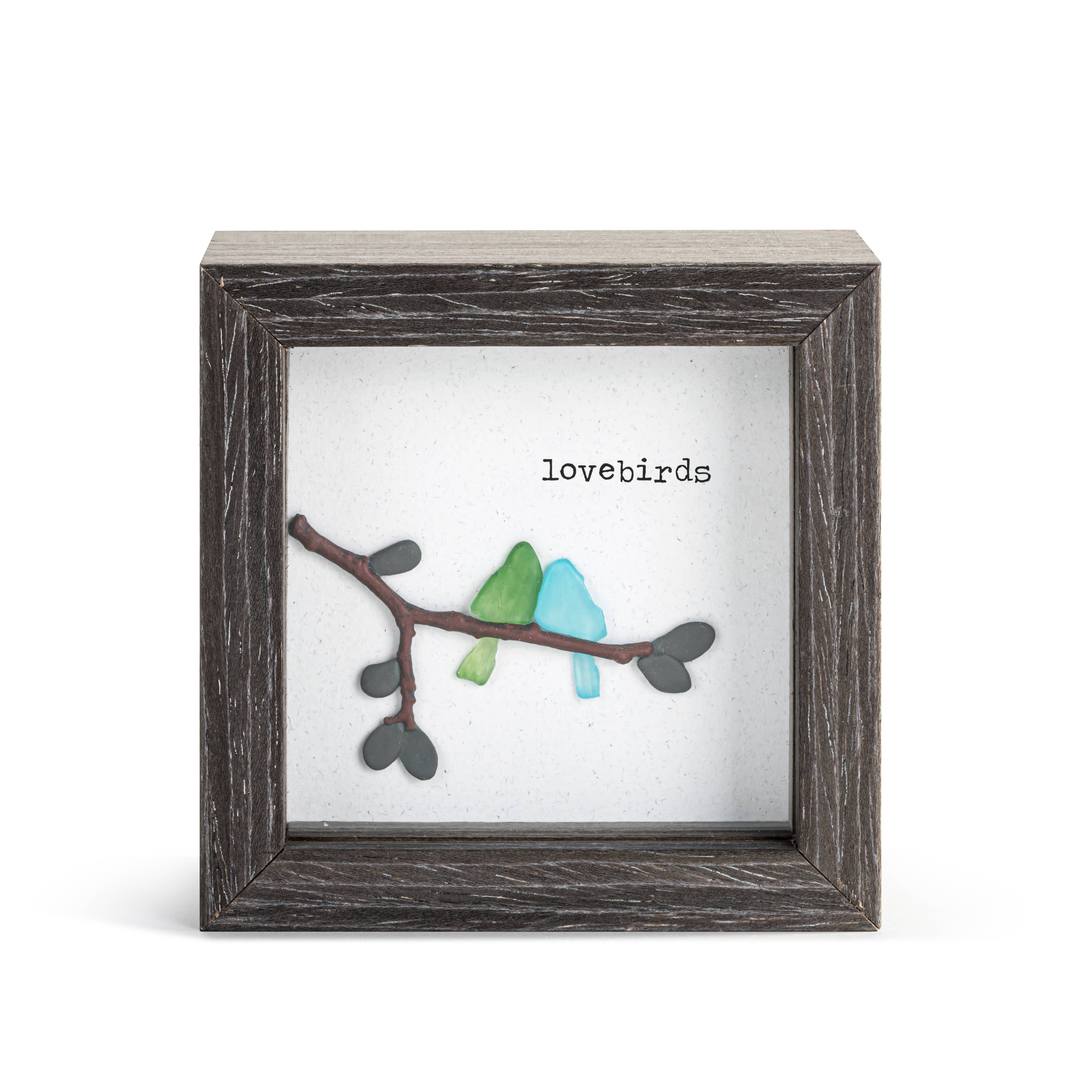 A square framed art piece featuring a rustic dark gray wood frame with a white backing. On the backing is a branch with heart-shaped leaves in dark gray and two small birds in light green and light blue, with the word 'lovebirds' printed above.