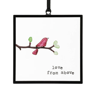 Love From Above Suncatcher by DEMDACO — a solitary crimson sea glass bird perched on a pebble branch with green sea glass leaf buds, inside a square black metal floating frame with "love from above" in typewriter font, hung by a black ribbon