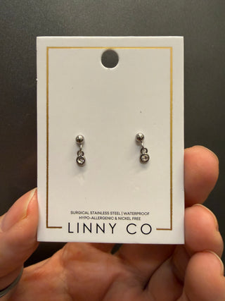 A pair of small silver drop earrings on a white display card with the brand name LINNY CO, held by a hand against a dark gray background.