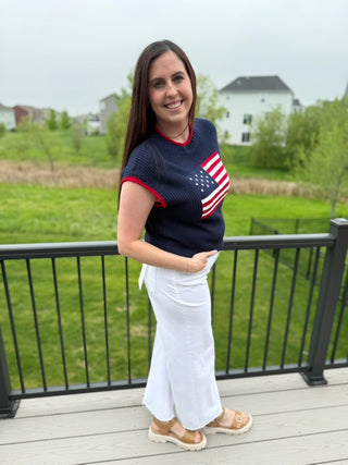 Woman wearing a navy sweater with an American flag design on a deck with a grassy field in the background.
