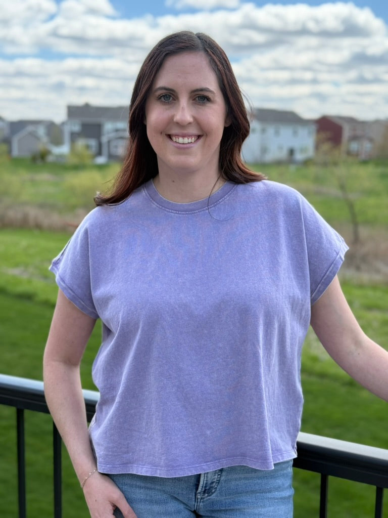 Woman wearing a purple t-shirt standing outdoors with houses in the background